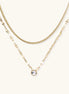 Layered gold necklace with herringbone and link chains, small bezel-set crystal pendant on cream background