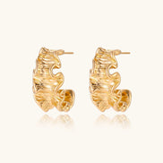 Pair of gold huggie hoops with sculpted wave texture and dimensional ripples on neutral background.