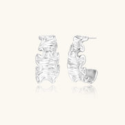 Textured white gold-plated hoop earrings with flowing wave pattern against cream background, showing dimensional rippled surface.