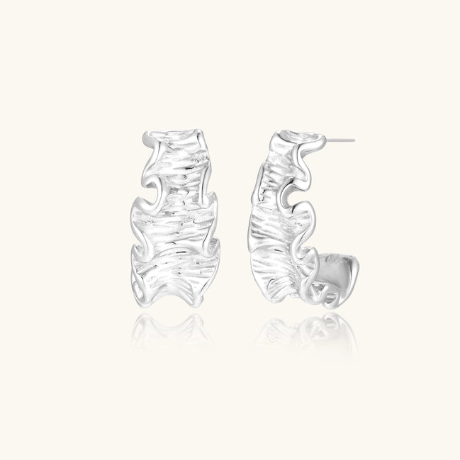 Textured white gold-plated hoop earrings with flowing wave pattern against cream background, showing dimensional rippled surface.