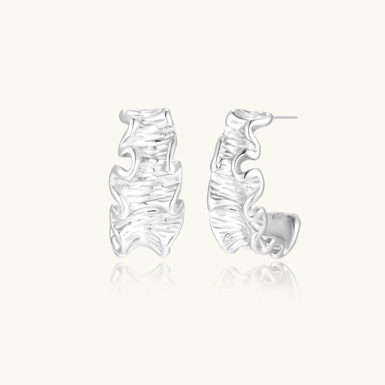 Textured white gold-plated hoop earrings with flowing wave pattern against cream background, showing dimensional rippled surface.