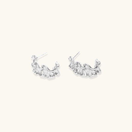 Textured white gold-plated hoop earrings with flowing wave pattern against cream background, showing dimensional rippled surface.