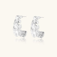 Textured white gold-plated hoop earrings with flowing wave pattern against cream background, showing dimensional rippled surface.