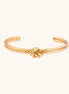 Gold cuff bracelet with twisted rope texture and decorative knot centerpiece on cream background.