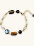 Gold chain bracelet with tiger's eye, obsidian, pearl, and iridescent abalone stones on cream background.